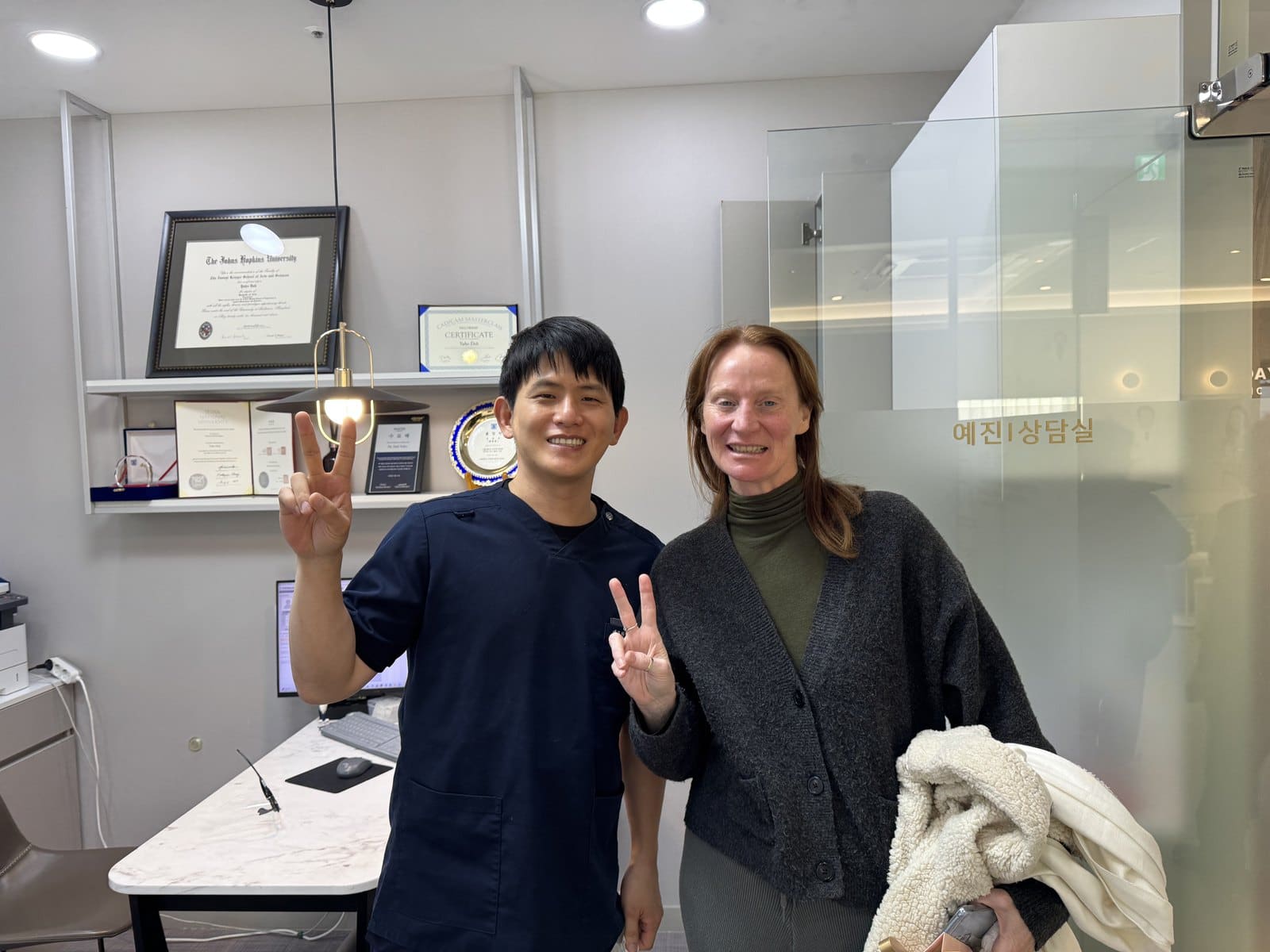 An international patient and Dr. Doh giving peace signs in front of the consultation room, with his framed Johns Hopkins diploma on the shelf