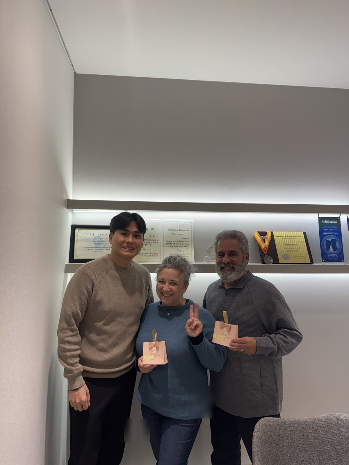 An international couple holding gift bags with Dr. Doh in front of the clinic's certificate shelf after finishing treatment